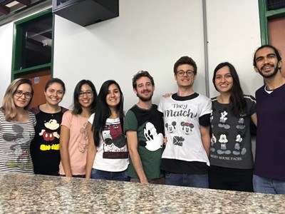 Foto de membros anteriores do LaGenBio no corredor do Departamento de Genética e Evolução da Ufscar. Na parte inferior podemos ver parte de uma bancada de mármore e atrás da mesma estão os membros Karen, Bruna, Fabrícia, Katiane, Djaci, Mateus, Maria e Thiago Machado. Todos estão vestindo camisetas com desenhos do Mickey ou Minie Mouse, exceto o Thiago que veste uma blusa azul arroxeado escuro. Ao fundo observamos a parede do prédio com duas portas de madeira e batentes verde. Em ordem podemos ver Bruna Martins (vestindo blusa preta com desenho do Mickey Mouse), Fabrícia 