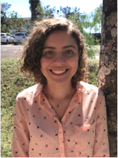 Aluna Bruna Martins Garcia. Mulher jovem de pele branca, apresenta cabelos cacheados pouco acima dos ombros e olhos verdes. Está usando uma camisa de cor salmão com pequenos desenhos de guarda-chuvas pretos por todo o tecido. Também está usando uma corrente prateada com pingente quadrado e sorri.
