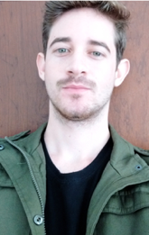 Aluno José Djaci Augusto Neto. Homem branco, jovem de olhos verdes e cabelo castanho médio e corte curto. Apresenta barba curta, castanha em formato de cavanhaque, veste uma camiseta preta com uma jaqueta verde militar por cima.