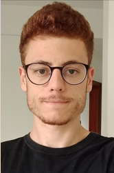 Mateus Priolo Grejo. Homem jovem ruivo de pele branca, cabelo avermelhado curto. Ele usa um óculos arredondado de armação preta e uma camiseta na cor preta.