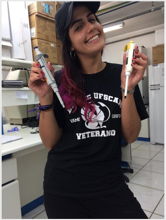 Aluna Rachel Marascalchi. Mulher jovem de pele branca bronzeada, olhos castanhos, cabelo castanho com pontas rosas. Ela usa um boné preto e roupas pretas, está sorrindo com a cabeça inclinada para a esquerda enquanto segura uma pipeta branca com detalhes amarelos com a mão esquerda e uma pipeta cinza com detalhes azuis na mão direita.