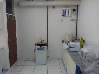 Imagem do Laboratório de biologia molecular, vista mais aproximada do final do laboratório a partir da porta de entrada. Possível verificar a parede do fundo com autoclave encostada, do lado esquerdo a porta de madeira que dá acesso ao laboratório de cultivo celular e manipulação genética. Do lado direito vemos o sistema de purificação de água, a pia com o barrilete de armazanamento de água deionizada, além da bancada por toda a extensão do lado direito com banho-maria sobre a mesma.