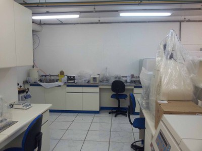 Imagem do Laboratório de biologia molecular, vista de dentro da segunda parte do laboratório. Do lado direito parte da bancada que divide o laboratório com a centrífuga, geldocumentador e computador sobre a mesma. Do lado esquerdo parte da bancada com balança simples e parte do armário fixado na parede. Centralizado na imagem está a parede do fundo com parte da pia e bancada com banho-maria, pHmetro e homogeneizador magnético.