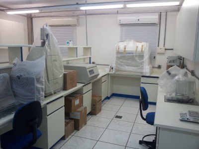 Imagem do Laboratório de biologia molecular, vista da entrada da segunda área do laboratório. Do lado esquerdo apresenta a bancada que separa o laboratório em 2 áreas, com computador, geldocumentador, e centrífuga refrigerada sobre a mesma. na parede do fundo observamos uma janela com persiana horizontal branca, condicionador da ar sobre a persiana e a frente da mesma uma capela de exaustão que fica apoiada em uma bancada que se conecta com outra parte que se estende por toda a extensão da parede do lado direito. Sobre a bancada do lado direito é possível observar homogeneizador de tecidos, eletrofusor, balança analítica e balança simples. Acima dos equipamentos podemos verificar um armário por toda extensão da parede do lado direito. 