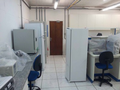 Imagem do Laboratório de biologia molecular, vista para a porta. Ao fundo e centralizado está a porta de entrada/saída do laboratório. Do lado direito observa-se parte da bancada que divide o laboratório com o computador e freezer vertical encostado na mesma. Do lado esquerdo verifica-se a bancada encostada na parede com banho-maria, pHmetro e estufa de esterilização. Encostado na bancada observamos parte da geladeira e do ultrafreezer.