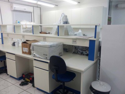 Imagem do Laboratório de biologia molecular, vista intermediária. Podemos observar a continuação da bancada que divide o laboratório, sendo de cor branca com detalhes azul. Sobre a mesma podemos observar a centrífuga e alguns equipamentos do outro lado do laboratório. Encostado no final dessa bancada podemos observar a traseira do freezer -20ºC e um tambor para coleta de lixo.